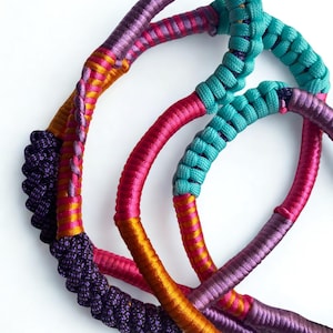 Puede incluir: Primer plano de varios collares de cuerda trenzada coloridos. Los collares presentan una variedad de colores, incluyendo morado, rosa, turquesa y dorado. La cuerda está tejida apretadamente, creando una superficie texturizada. Los collares están dispuestos en un patrón circular.
