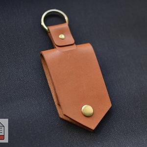 Puede incluir: Un llavero de cuero marrón con un llavero dorado y un cierre de botón a presión de latón. El soporte es rectangular con un diseño plegado y una base puntiaguda. El fondo es de tela azul oscuro.