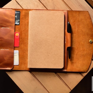 May include: A brown leather notebook cover with a pen holder, card slots, and a notepad. The cover has a stitched design and a brass button.