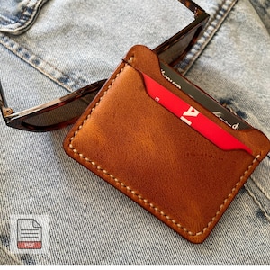 May include: A brown leather cardholder with white stitching and a red card partially visible. Tortoiseshell sunglasses rest on the cardholder. The cardholder is on a denim surface.