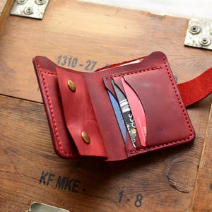 May include: Open red leather wallet with multiple card slots and a snap closure. The wallet is displayed open, revealing the interior compartments and cards. The background is a wooden surface.