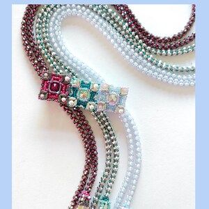 Może przedstawiać: Wielorzędowy naszyjnik z kwadratowym wisiorem ozdobionym różowym, turkusowym i przezroczystym kryształem. Naszyjnik wykonany jest z srebrnych, turkusowych i różowych koralików. Naszyjnik nazywa się "Tempesta Necklace" autorstwa Carmelina Calabrese.