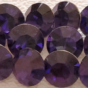 Puede incluir: Un grupo de 12 piedras de cristal redondas y facetadas de color morado. Las piedras de cristal están dispuestas en forma de semicírculo.