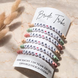Könnte beinhalten: Sechs Perlenarmbänder mit den Worten "Team Bride" und einem kleinen roten Herzen auf jedem Armband. Die Armbänder sind übereinander auf einer weißen Karte mit den Worten "Bride Tribe" und "Eine Nacht. Ein Team. Ein Ziel. Unvergesslich feiern!" in schwarzer Schrift gestapelt.