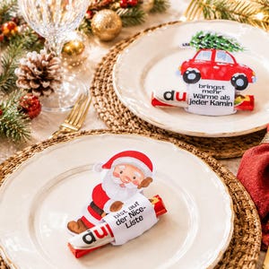 May include: Two chocolate bar wrappers featuring Christmas designs on white plates. One wrapper shows a red car with a tree and text reading bringst mehr Wärme als jeder Kamin. The other shows Santa Claus with text reading bist auf der Nice-Liste.