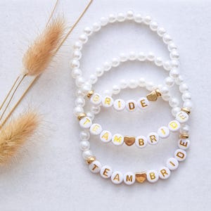 Könnte beinhalten: Drei weiße Perlenarmbänder mit goldenen Akzenten und weißen Buchstabenperlen mit der Aufschrift "BRIDE" und "TEAM BRIDE". Die Armbänder haben goldfarbene herzförmige Anhänger. Die Armbänder liegen auf einer weißen Oberfläche.