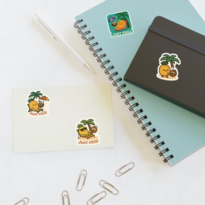 May include: A collection of stickers featuring a cartoon pineapple character relaxing under a palm tree with a coconut. The stickers have the text "Just Chill" and are on a light blue notebook, a black notebook, and a cream-colored card.