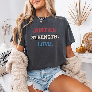 Könnte beinhalten: Dunkelgraues T-Shirt mit den Worten "JUSTICE. STRENGTH. LOVE." in roter, weißer und blauer Schrift. Das Shirt wird mit Jeansshorts und einem cremefarbenen Pullover getragen. Eine silberne Halskette ist sichtbar.