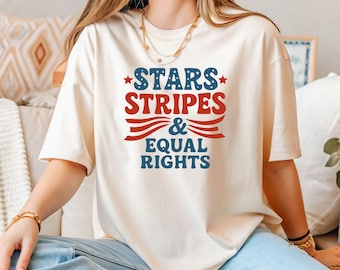 May include: Cream-colored t-shirt with the text "STARS STRIPES & EQUAL RIGHTS" in red and blue. The design includes stars and stripes, evoking a patriotic theme. The shirt is a casual, short-sleeved style.