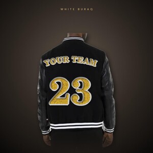 Può includere: Una giacca varsity nera a maniche lunghe, con la scritta WHITE BURAQ in alto. Il retro mostra le parole YOUR TEAM in lettere dorate sopra il grande numero 23 dorato. La giacca presenta bordi a coste a righe bianche e nere.