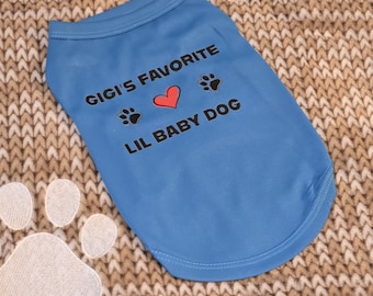 May include: A light blue dog t-shirt with the text "GIGI'S FAVORITE LIL BABY DOG" in black, a red heart, and paw prints. The shirt is on a brown and beige knitted surface. A white paw print is in the lower left corner.