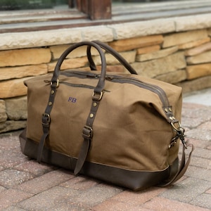 May include: A brown canvas duffel bag with leather accents. The bag features a top zipper, two handles, and a detachable shoulder strap. The initials "FB" are embroidered on the front. Metal hardware and leather straps secure the bag's design.