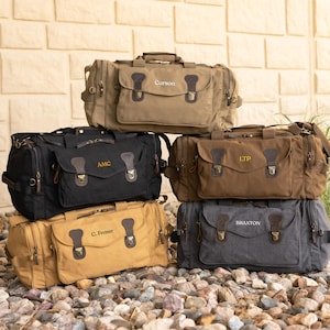May include: A stack of six canvas duffel bags in various colors: olive green, black, brown, tan, and gray. Each bag features leather accents, metal hardware, and personalized text. The bags are sitting on a bed of small rocks.