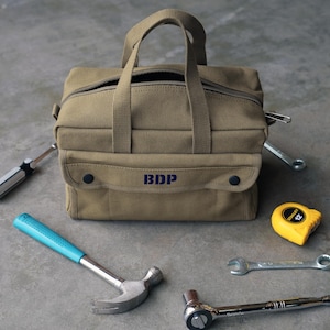 May include: A tan canvas tool bag with a zipper closure and a handle, embroidered with the letters "BDP". The bag is surrounded by tools, including a hammer, screwdrivers, wrenches, and a tape measure.