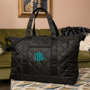 May include: Black quilted tote bag with teal monogram "SBG". The bag has a diamond pattern, a zippered front pocket, and two top handles. It also includes a detachable shoulder strap. The bag is sitting on a patterned ottoman.