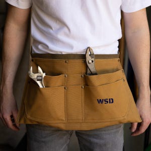 May include: A tan canvas tool belt with multiple pockets, riveted accents, and a dark green belt. A wrench and pliers are visible in the pockets. The initials "WSD" are printed in navy blue on the front.