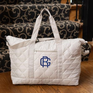 May include: A cream-colored quilted tote bag with a navy blue monogram. The bag has a zippered front pocket, a shoulder strap, and two top handles. The bag is sitting on a wooden surface.