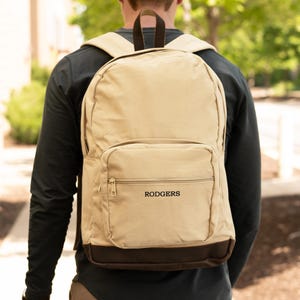 May include: Tan backpack with a brown base and straps. The backpack has a front zippered pocket and the name "RODGERS" printed in black. The backpack is being worn by a person.