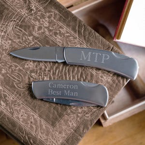 May include: Two silver pocket knives with engraved text. The top knife has the initials "MTP" engraved on the handle. The bottom knife has the text "Cameron Best Man" engraved on the handle.