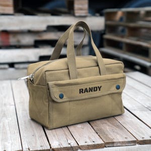 May include: A tan canvas tool bag with two top handles and a zippered main compartment. The front features a flap pocket secured by two dark blue snaps, with the name RANDY printed in black capital letters on the center of the pocket flap.