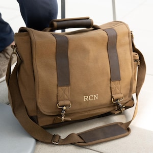 May include: A brown canvas messenger bag with leather accents. The bag features a top handle, shoulder strap, and a flap closure. The initials "RCN" are embroidered on the front. The bag has metal hardware.