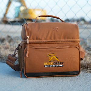 May include: Brown insulated lunch bag with a top handle, shoulder strap, and multiple compartments. Features the "Buildcraft Construction" logo in yellow and white. Includes a side mesh pocket and a zipper closure. The bag has a brown exterior with a black trim.