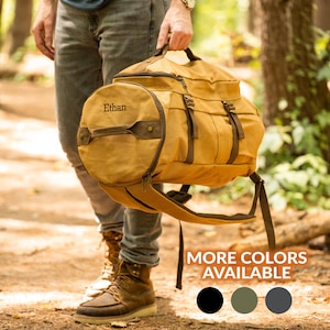 May include: A mustard-colored duffel bag with brown accents and straps. The bag has a cylindrical end with the name "Ethan" embroidered on it. The bag is being held by a person in a natural outdoor setting. The text "MORE COLORS AVAILABLE" is visible.