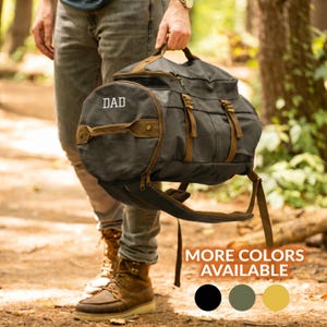 May include: A charcoal grey canvas duffel bag with brown leather-like accents, featuring the word DAD embroidered in white on the circular side panel. The bag includes brown straps, metal buckles, and a shoulder strap, with text stating MORE COLORS AVAILABLE.