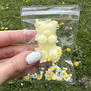 May include: A clear plastic bag filled with yellow and pastel-colored confetti and a yellow gummy bear. The gummy bear has a rounded shape with small yellow embellishments. The confetti includes stars, moons, and small yellow spheres.
