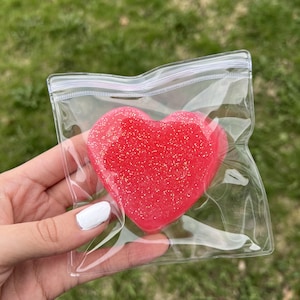 May include: A red heart-shaped item with glitter is enclosed in a clear plastic bag. The bag is sealed with a zipper closure. The background is blurred green grass.
