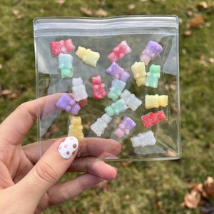 May include: A clear plastic bag filled with colorful gummy bear-shaped candies. The candies are in various colors, including red, yellow, purple, and green. The bag is held in a person's hand, with a blurred green background.
