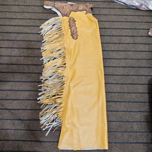 Peut inclure: Chaps en cuir jaune moutarde avec de longues franges sur les côtés. Les chaps ont un empiècement décoratif en cuir marron avec des motifs floraux et un écusson décoratif assorti. Conçu pour l'équitation.