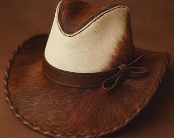 May include: A cowboy hat with a brown and white cowhide pattern. The hat has a brown leather band with a bow detail and a stitched brim. The hat is a classic Western style.