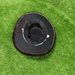Pode incluir: Um chap&eacute;u preto com acabamento em pele castanha e uma correia de queixo em couro castanho. O interior do chap&eacute;u &eacute; preto com uma faixa castanha. O chap&eacute;u est&aacute; sobre uma superf&iacute;cie verde.
