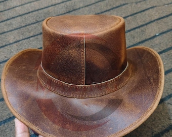 Puede incluir: Sombrero de vaquero de cuero marrón con ala ancha y corona cilíndrica. El sombrero tiene un aspecto desgastado, con desgaste visible, y lo sostiene una mano. El color del sombrero es un marrón rico y terroso.
