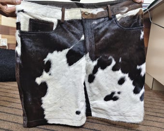 May include: Cowhide shorts with a brown leather belt and pockets. The shorts feature a black and white spotted pattern. The shorts are held up by a person's hands.