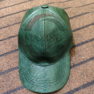 May include: Green baseball cap with a textured, crocodile-like pattern. The cap has a curved brim and a button on top. The letters "MA" are visible on the front of the cap.