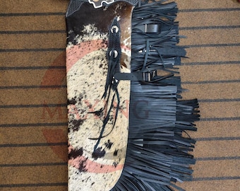 Könnte beinhalten: Cowhide-Chaps mit schwarzem Lederfransen. Die Chaps haben ein Kuhfell-Panel mit braunen, schwarzen und rosa Markierungen. Der obere Teil ist schwarz mit einer gewebten Textur und einem Schnallenverschluss. Ein dekoratives Emblem ist angebracht.