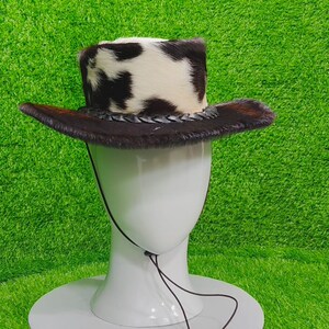Pode incluir: Um chap&eacute;u de cowboy com uma coroa de pele de vaca e uma aba de pele castanha. O chap&eacute;u &eacute; exibido em uma cabe&ccedil;a de manequim branca sobre um fundo verde. O chap&eacute;u tem uma faixa de couro tran&ccedil;ado.