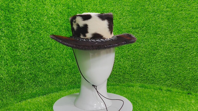 Pode incluir: Um chap&eacute;u de cowboy com uma coroa de pele de vaca e uma aba de pele castanha. O chap&eacute;u &eacute; exibido em uma cabe&ccedil;a de manequim branca sobre um fundo verde. O chap&eacute;u tem uma faixa de couro tran&ccedil;ado.