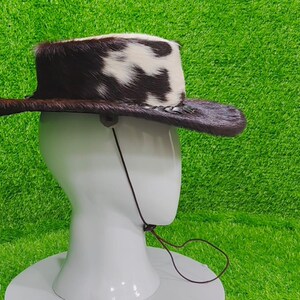Pode incluir: Um chap&eacute;u de cowboy com aba de couro marrom e coroa coberta com uma mistura de pele de vaca marrom e branca. O chap&eacute;u tem uma tira de queixo e &eacute; exibido em uma cabe&ccedil;a de manequim branca. Acess&oacute;rio de estilo ocidental.