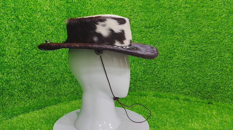 Pode incluir: Um chap&eacute;u de cowboy com aba de couro marrom e coroa coberta com uma mistura de pele de vaca marrom e branca. O chap&eacute;u tem uma tira de queixo e &eacute; exibido em uma cabe&ccedil;a de manequim branca. Acess&oacute;rio de estilo ocidental.