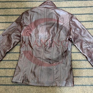 May include: A brown leather jacket with a collared neckline and long sleeves. The jacket has a tailored fit and visible stitching details. The back of the jacket is visible, showing vertical seams and a classic design.