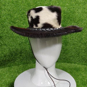 Pode incluir: Chap&eacute;u de cowboy com coroa manchada em preto e branco e aba marrom escuro. O chap&eacute;u apresenta uma faixa de couro tran&ccedil;ado e uma tira de queixo. O chap&eacute;u &eacute; exibido em uma cabe&ccedil;a de manequim branca sobre um fundo verde.