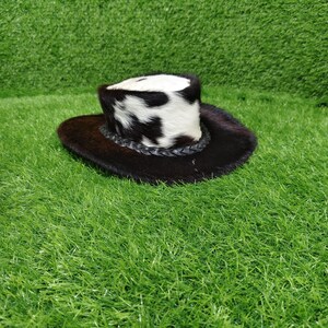 Pode incluir: Um pequeno chap&eacute;u de cowboy com aba marrom escuro e copa manchada em preto e branco. O chap&eacute;u apresenta uma faixa de couro tran&ccedil;ado. O chap&eacute;u &eacute; feito de um material semelhante a pele.