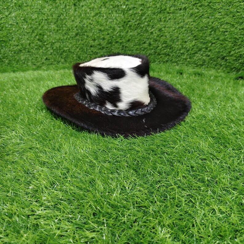 Pode incluir: Um pequeno chap&eacute;u de cowboy com aba marrom escuro e copa manchada em preto e branco. O chap&eacute;u apresenta uma faixa de couro tran&ccedil;ado. O chap&eacute;u &eacute; feito de um material semelhante a pele.
