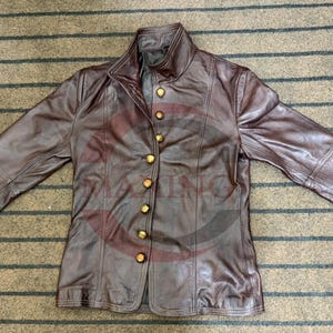 May include: A brown leather jacket with a high collar and gold-colored buttons. The jacket has long sleeves and a tailored fit. The leather appears worn, giving it a vintage look. The jacket is laid flat on a striped surface.