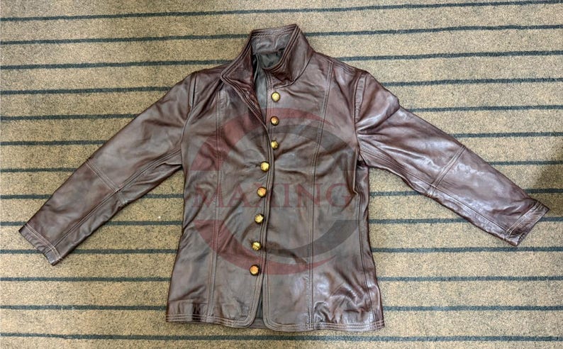 May include: A brown leather jacket with a high collar and gold-colored buttons. The jacket has long sleeves and a tailored fit. The leather appears worn, giving it a vintage look. The jacket is laid flat on a striped surface.