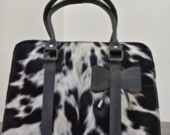 May include: A rectangular handbag with a black and white cowhide pattern. The bag has two black handles and a decorative bow on the front. The handles and bow are made of dark gray leather. The bag has silver-colored hardware.