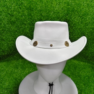 May include: A white cowboy hat with a wide brim and a rounded crown. The hat features a decorative band with two circular, gold-colored embellishments and a small, centered stud. The hat is displayed on a white mannequin head against a green background.